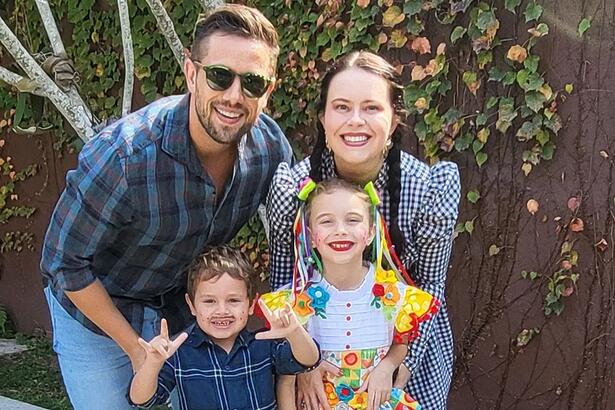 Mariana Bridi com vestido quadriculado e calça jeans, sorrindo, com tranças no cabelo; Rafael Cardoso com camisa quadriculada em tons de azul e branco, sorrindo, de oculos de sol, segurando os filhos durante passeio