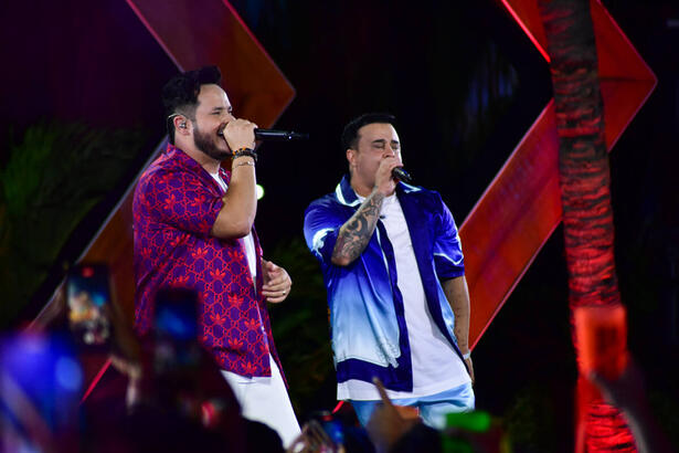 Matheus, de camisa em tom de magenta, estampada, segurando o microfone com a mão direita, ao lado de Kauan, de camisa em degrade azul e branco, durante gravação de EP