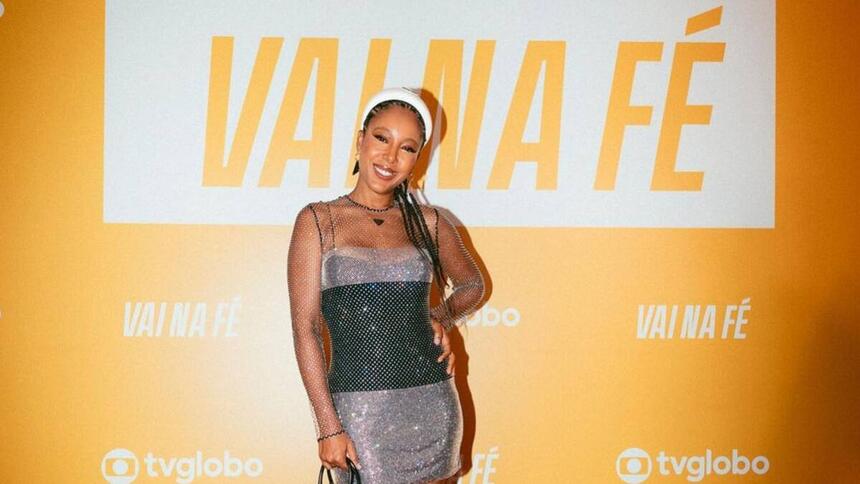 Cantora Negra Li com vestido de brilho cinza com preto sorrindo e apoiando a mão na cintura, com fundo de lançamento amarelo da novela 'Vai Na Fé' da TV Globo.