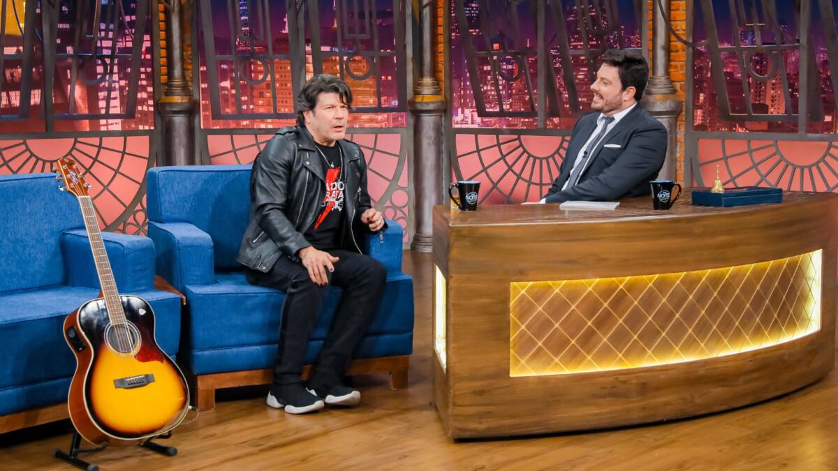 Cantor Paulo Ricardo de roupa preta sentado no sofá azul do The Noite olhando para Danilo Gentili, apresentador do programa que está apoiado na bancada e vestindo terno com gravata azul.