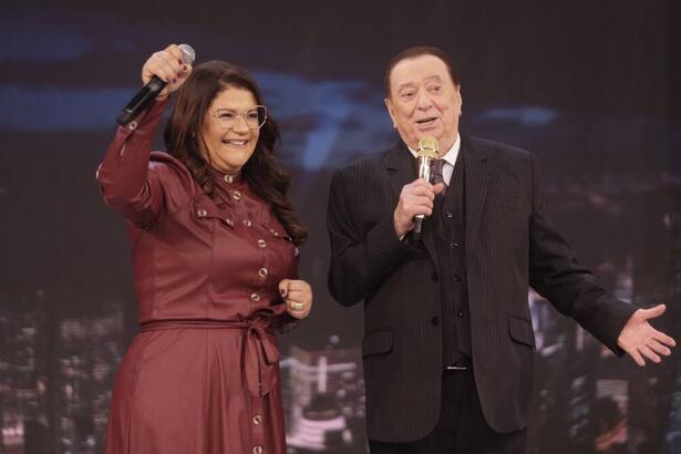 Midian Lima vestindo uma roupa vinho, segurando o microfone e sorriso. Ao lado, Raul Gil de terno preto com a mão ao lado e uma segurando o microfone.