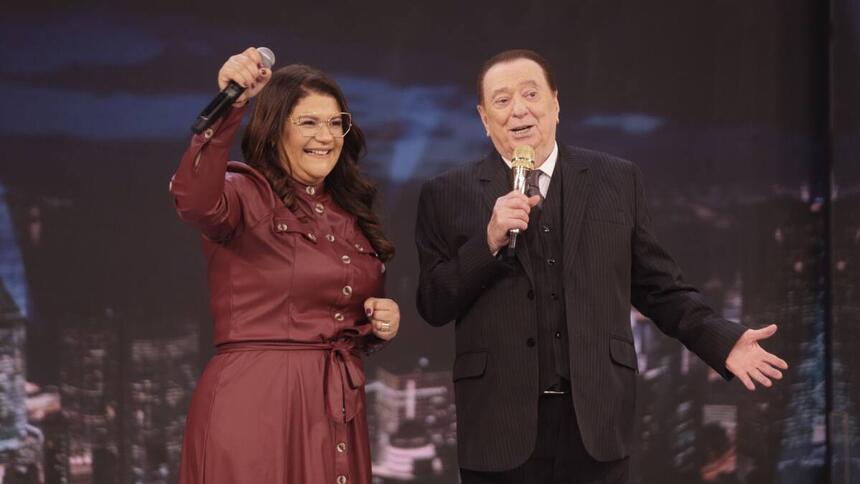 Midian Lima vestindo uma roupa vinho, segurando o microfone e sorriso. Ao lado, Raul Gil de terno preto com a mão ao lado e uma segurando o microfone.