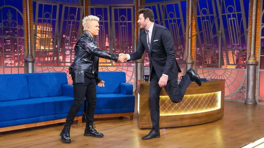 Billy Idol convidado do The Noite vestido e roupa preta e cabelo loiro, cumprimenta Danilo Gentili que está vestido de terno preto e perna cruzada.