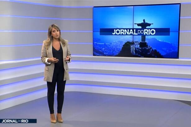 Apresentadora do Jornal do Rio na Band do Rio de Janeiro no estúdio do programa vestida com macacão preto e um casaco da cor marron.