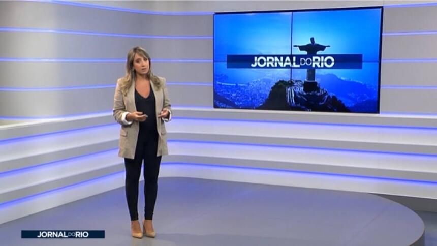 Apresentadora do Jornal do Rio na Band do Rio de Janeiro no estúdio do programa vestida com macacão preto e um casaco da cor marron.