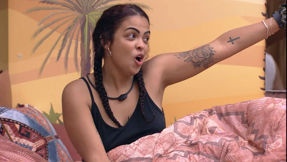 Paula Freitas de top preto, de trança, sentada na cama do Quarto Deserto, do BBB 23