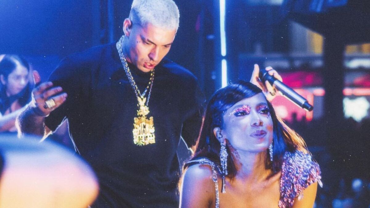 Filipe Ret de camisa preta e correntes no pescoço segura o microfone olhando para Anitta. Cantora está fazendo biquinho com a boca e mãos no joelhos durante um apresentação de um show.