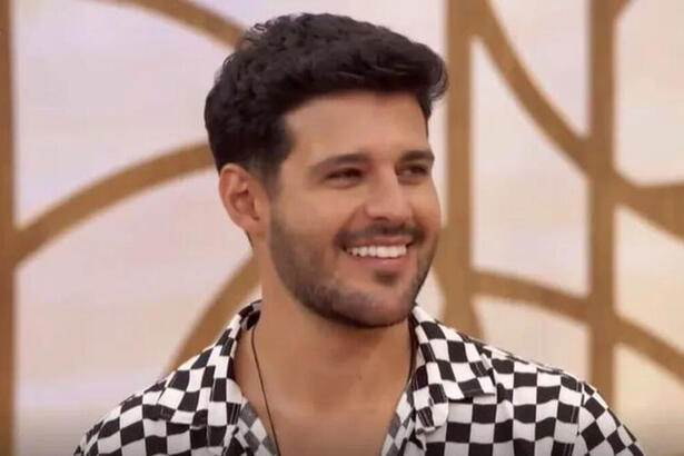 Rodrigo Mussi com uma roupa xadrez em preto e branco, sorriso aberto. Ele está sentado no sofá do Encontro