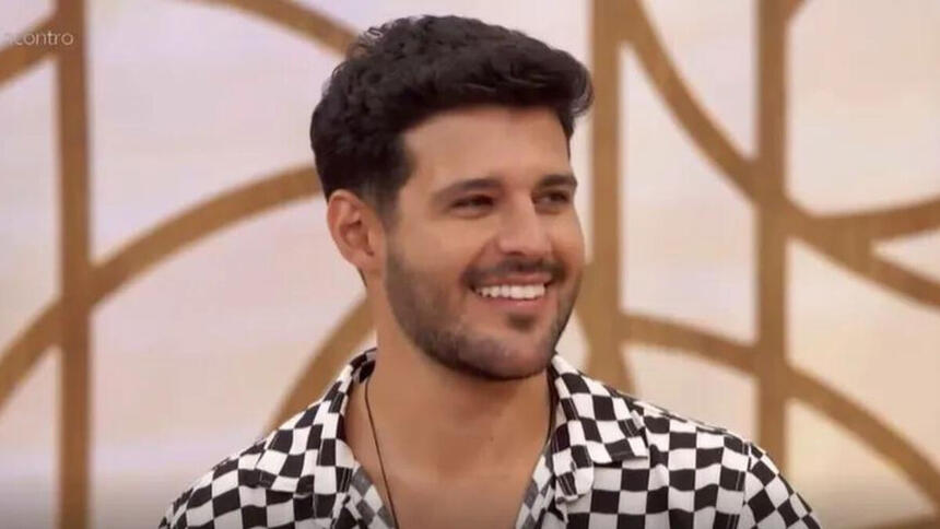 Rodrigo Mussi com uma roupa xadrez em preto e branco, sorriso aberto. Ele está sentado no sofá do Encontro