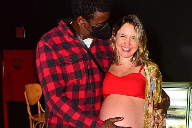 Seu Jorge de camisa quadriculada vermelha e preta, de mascara preta, com a mao esquerda na barriga de Karina durante evento em São Paulo