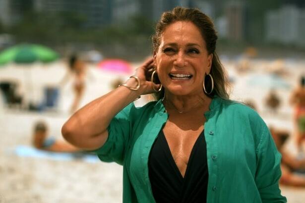 Susana Vieira de camisa preta e verde, com a mão na cabeça, sorrindo, durante gravação externa na praia