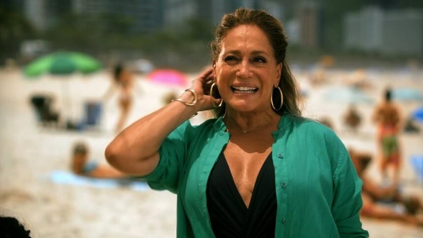 Susana Vieira de camisa preta e verde, com a mão na cabeça, sorrindo, durante gravação externa na praia