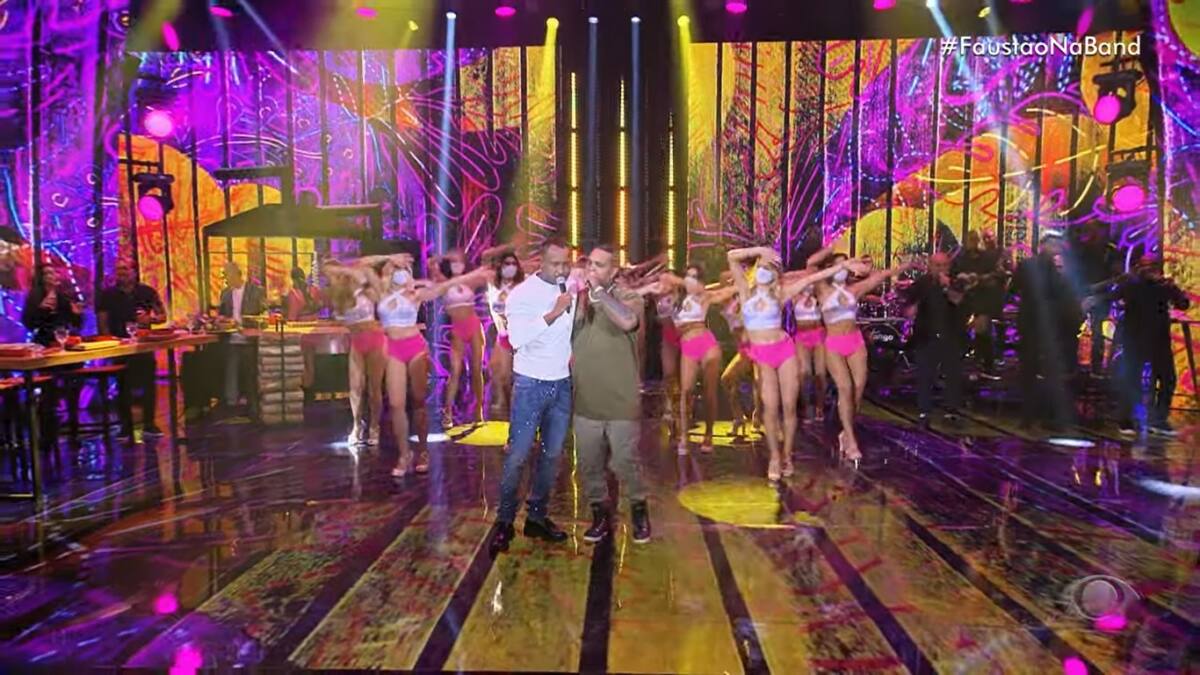 Thiaguinho de camisa branca e calça azul ao lado de Rodriguinho com roupa marrom no palco do Faustão Na Band. Também estão as bailarinas que vestem camisa branca e saia rosa.