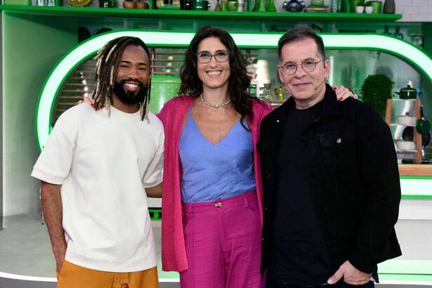 João Diamente de camisa branca, Paola Carosella com camisa azul, casaco e calça rosa ao lado de Leandro Hassum vestido de preto no palco do novo reality da TV Globo ‘Minha Mãe Cozinha Melhor Que a Sua’.