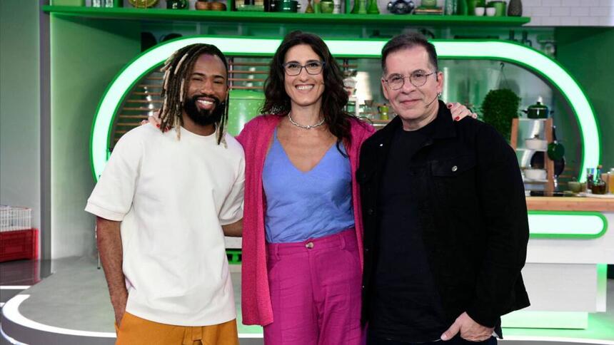 João Diamente de camisa branca, Paola Carosella com camisa azul, casaco e calça rosa ao lado de Leandro Hassum vestido de preto no palco do novo reality da TV Globo ‘Minha Mãe Cozinha Melhor Que a Sua’.