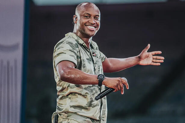 Thiaguinho sorrindo, com camisa de estampa camuflada, segurando o microfone com a mão direita durante gravação de DVD no Maracanã