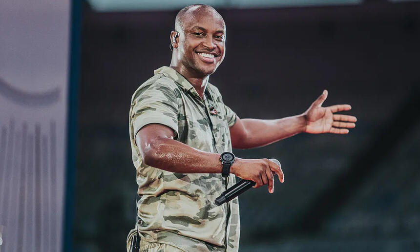 Thiaguinho sorrindo, com camisa de estampa camuflada, segurando o microfone com a mão direita durante gravação de DVD no Maracanã