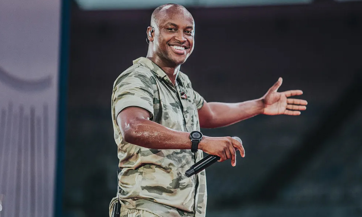 Thiaguinho sorrindo, com camisa de estampa camuflada, segurando o microfone com a mão direita durante gravação de DVD no Maracanã