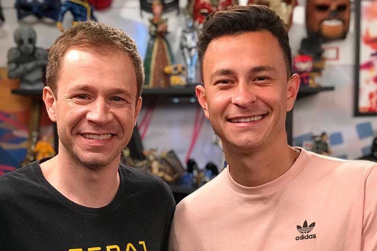 Tiago Leifert de camisa preta com estampa amarela, sorrindo, ao lado de Fred durante gravação de programa de game da TV Globo