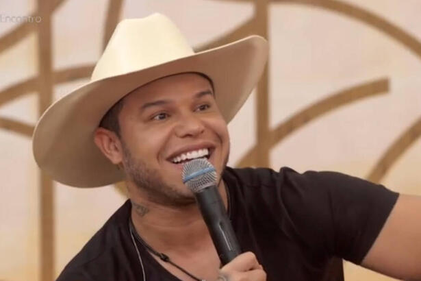 Tierry, com um chpéu branco na cabeça, sorrindo, com um microfone nas mãos, sentando no sofá do programa Encontro