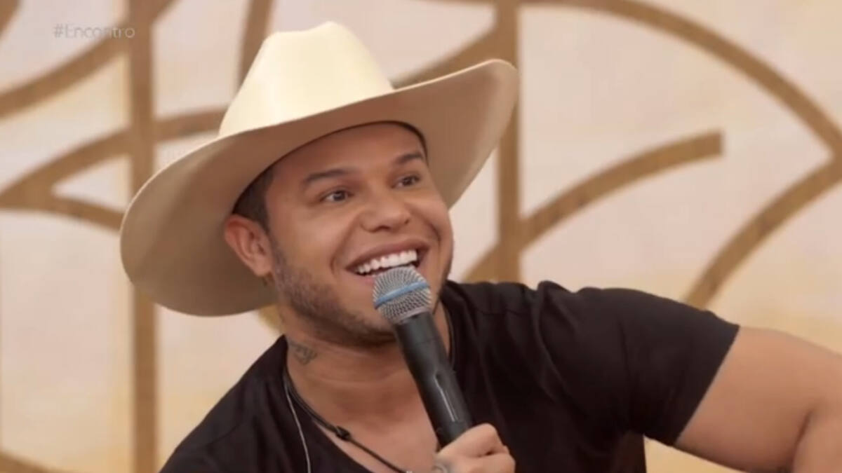 Tierry, com um chpéu branco na cabeça, sorrindo, com um microfone nas mãos, sentando no sofá do programa Encontro