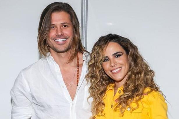 Wanessa Camargo de cabelo cacheado, sorrindo, de camisa social amarela, ao lado de Dado Dolabella, que está de camisa social branca, sorrindo, de cabelo liso e solto