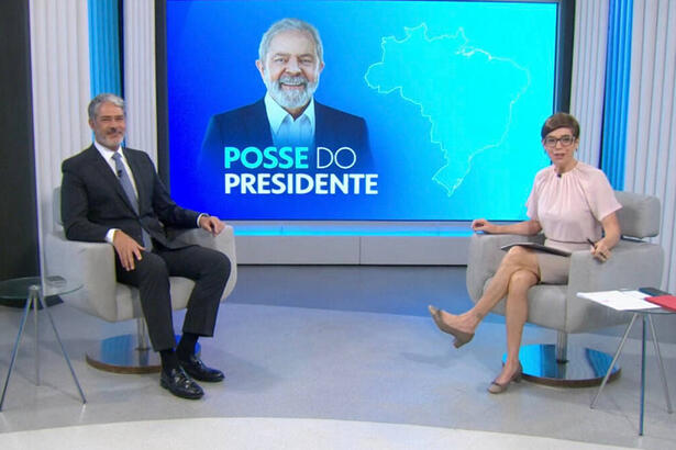 William Bonner e Renata Lo Prete sentados em poltranas, no estúdio da Globo em Brasília, em frente a um telão com imagem de Lula estampada