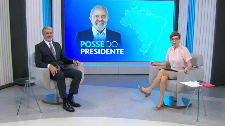 William Bonner e Renata Lo Prete sentados em poltranas, no estúdio da Globo em Brasília, em frente a um telão com imagem de Lula estampada