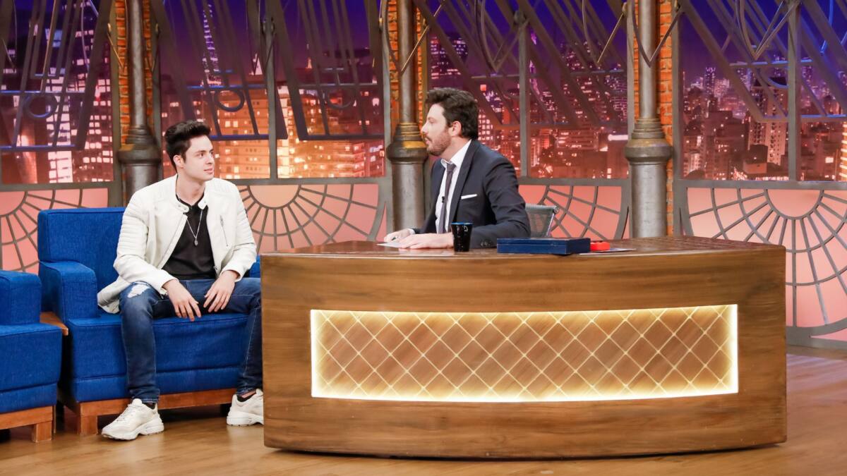 Enaldinho vestido de camisa preta e casaco branco sentando no sofá azul do The Noite. Ao lado está Danilo Gentili apresentador do programa apoiado na bancada vestindo terno preto.