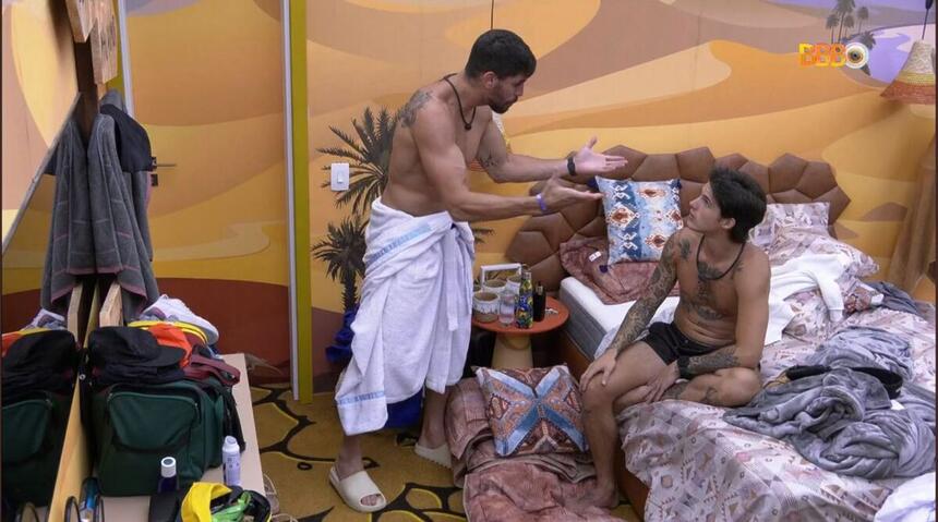 Cara de Sapato em pé no quarto do BBB 23, vestido com um roupão. Gabriel sentado na cama de frente para Cara de Sapato.