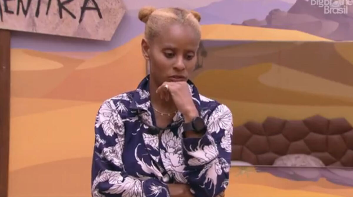 Aline Wirley com a mão no queixo, de camisa social branca com azul, cabelo preso, em pé, no Quarto Deserto, do BBB 23