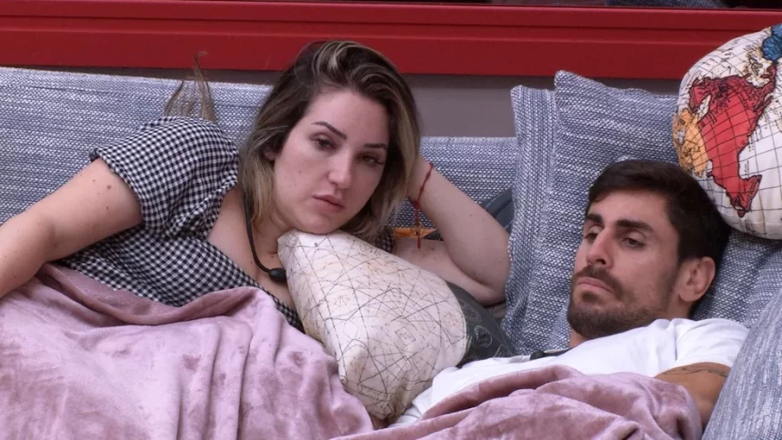 Amanda de cabelo solto, mão esquerda na cabeça, deitada ao lado de Cara de Sapato no sofá da sala do BBB 23