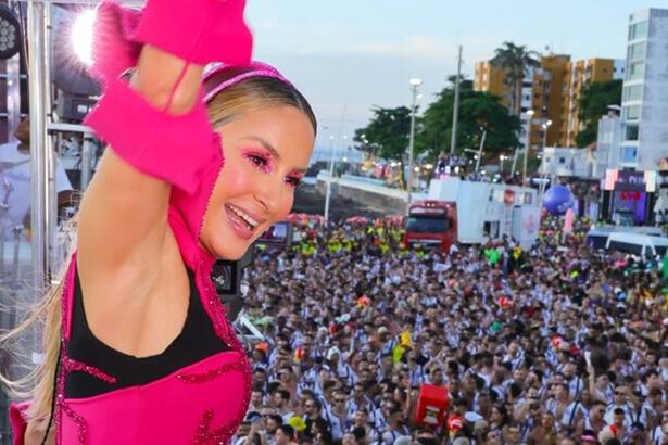 Claudia Leitte de look rosa com o braço pra cima, sorrindo, no trio em salvador