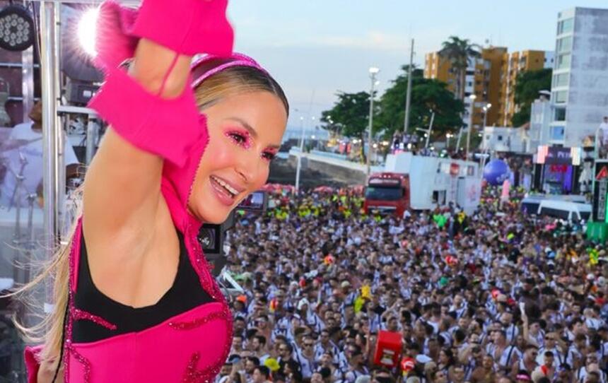 Claudia Leitte de look rosa com o braço pra cima, sorrindo, no trio em salvador