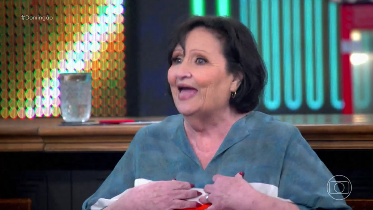 Dona Dea de vestido verde, com as mãos na caixa do peito, sentada durante pariticpação no Domingão, da TV Globo