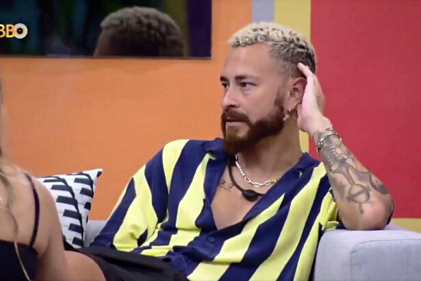 Fred de camisa listrada em amarelo e azul, com a mão esquerda a cabeça, sentado num sofá, na varanda do BBB