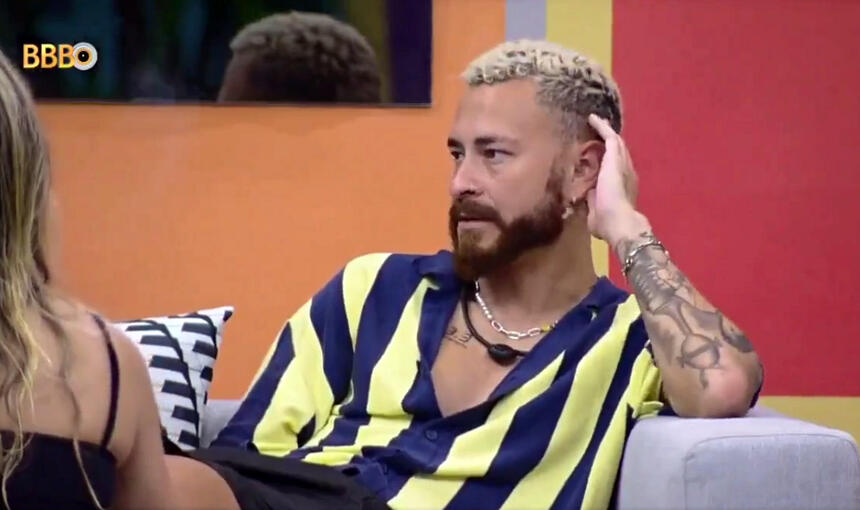 Fred de camisa listrada em amarelo e azul, com a mão esquerda a cabeça, sentado num sofá, na varanda do BBB