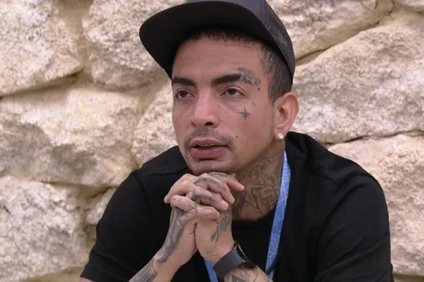 MC Guimê de camisa e bone preto, com colar azul e mãos juntas, sentado no banheiro do BBB 23