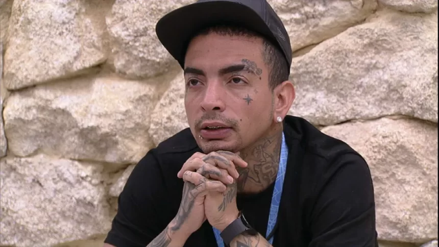 MC Guimê de camisa e bone preto, com colar azul e mãos juntas, sentado no banheiro do BBB 23