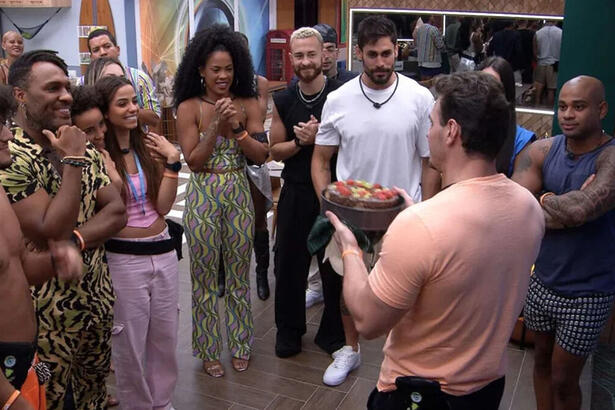 Gustavo, de costas, com o bolo nas mãos, na frente dos demais brothers na cozinha do BBB 23