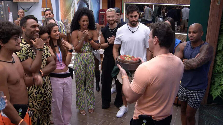 Gustavo, de costas, com o bolo nas mãos, na frente dos demais brothers na cozinha do BBB 23