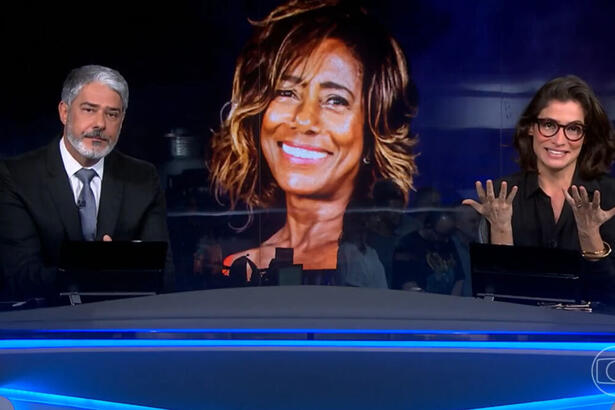 William Bonner e Renata Vasconcellos, na bancada do Jornal Nacional, com a imagem de Glória Maria ao fundo no telão da redação