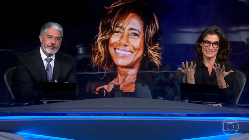 William Bonner e Renata Vasconcellos, na bancada do Jornal Nacional, com a imagem de Glória Maria ao fundo no telão da redação