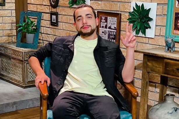 Lucas Selfie sentado com camisa amarela clara, jaqueta jeans preta, fazendo um 2 com a mão esquerda, com semblante sério nos estúdios de A Fazenda, da Record TV