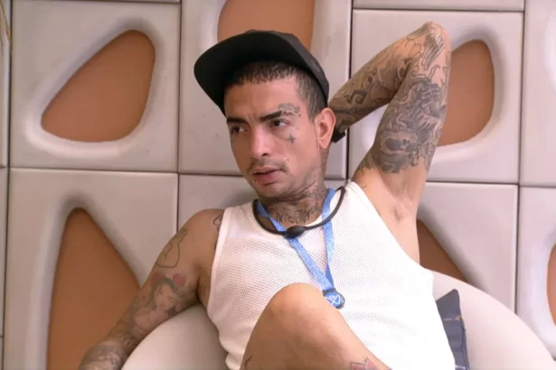 MC Guimê de camisa reggata branca, chapéu preto, colar azul sentado na varanda do BBB 23