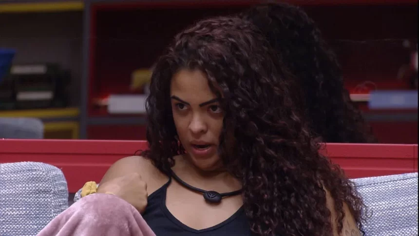 Paula Freitas de cabelo solto, camisa regata preta, microfone no pescoço, sentada no sofá da sala do BBB 23