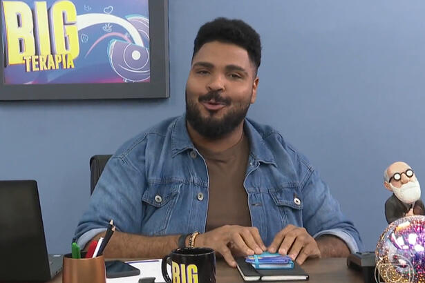 Paulo Vieira de camisa jeans, sentado em frente a uma bancada com adereços, um quadro, num estúdio azul gravando o Big Terapia, do BBB 23