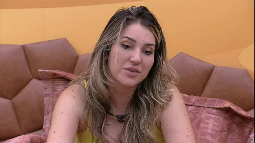 Amanda de regata amarela, cabelo solto, sentada na cama do Quarto Deserto do BBB 23