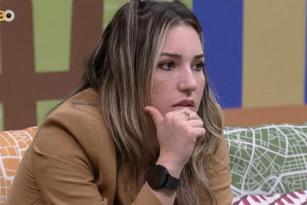 Amanda sentada nos sofás da área externa do BBB 23, com a mão no rosto e reflexiva