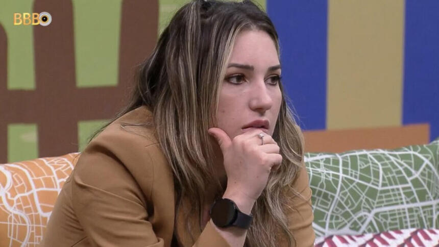 Amanda sentada nos sofás da área externa do BBB 23, com a mão no rosto e reflexiva